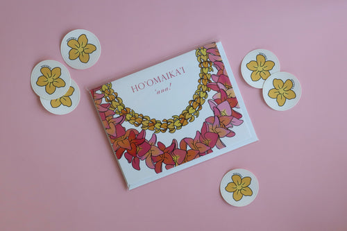Pink greeting card with floral Hawaiian lei art on the front and “ho’omaika’i ’Ana”, or “congratulations”. Flowers are puakenikeni and plumeria